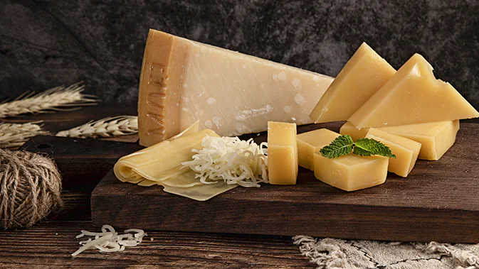 Zu sehen ist ein dunkles Holzbrett, auf dem verschiedene Käsesorten ansehnlich platziert sind. Es handelt sich dabei ausschließlich um Hartkäsesorten. Neben den Käsestücken ist auch etwas geriebener Käse auf dem Brett platziert.