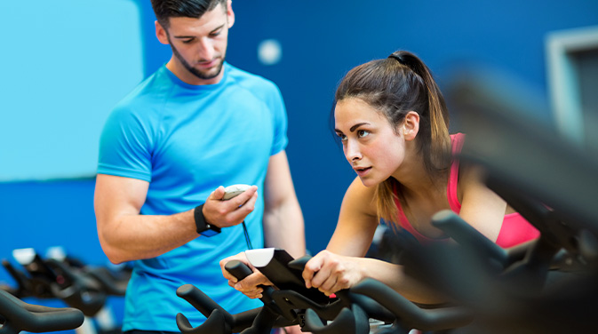 Trainer mit Stoppuhr neben einer Frau auf dem Ergometer