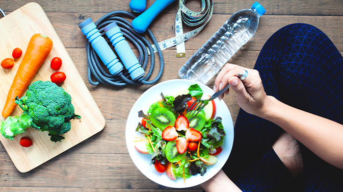 Frau sitzt auf dem Boden, mit einem Salat, umgeben von Sport-Tools