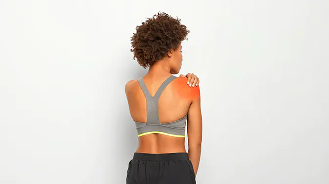 Afroamerikanische Frau von der Rückseite in Sportklamotten. Sie greift mit einer Hand an eine schmerzende Stelle an ihrer Schulter. Die schmerzende Stelle ist rot hervorgehoben.