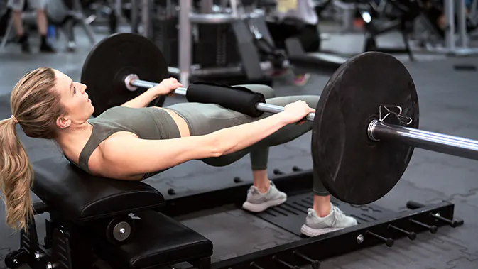 Eine Frau im Fitnessstudio führt die Übung Hip Thrusts mit einer Langhantel durch. Sie befindet sich gerade in der Position, in der die Hüfte nach oben durchgedrückt wird und der Oberkörper und die Oberschenkel eine gerade Linie bilden. 