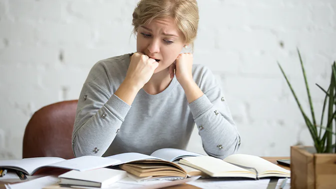 Eine Frau sitzt am Schreibtisch, vor ihr sind viele Bücher ausgebreitet und aufgeschlagen. Sie wirkt besorgt und ängstlich und beißt sich in die eigene Faust. 