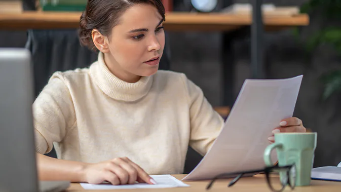 Eine junge Frau in hellem Rollkragenpullover  sitzt am Schreibtisch und schaut auf ein Blatt Papier mit Text. Sie ist offensichtlich am Lernen. Auf ihrem Tisch liegt eine Brille und eine Kaffeetasse.