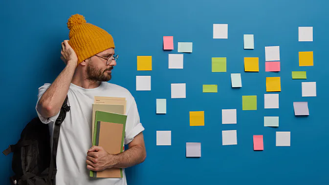 Ein Mann in einem weißen T-Shirt mit gelber Mütze, Brille und schwarzem Rucksack steht vor einer blauen Wand. An der Wand sind verschieden-farbige Post-its angebracht. Der Mann schaut auf die Zettel und kratzt sich am Kopf.