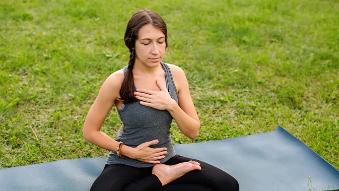 Eine Frau sitzt auf einer Yogamatte, die auf einer Wiese liegt. Sie sitzt im Schneidersitz und hat ihre Haare zu einem seitlichen Zopf geflochten. Eine Hand hat sie auf ihren Bauch gelegt, die andere auf ihren Brustkorb.