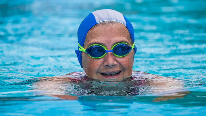 Eine ältere Dame mit Schwimmbrille und Badekappe schwimmt in einem Schwimmbecken. 