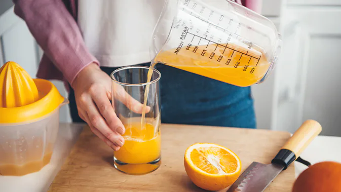 Eine Person schüttet frisch gepressten Orangensaft aus einem Messbecher in ein Glas. 