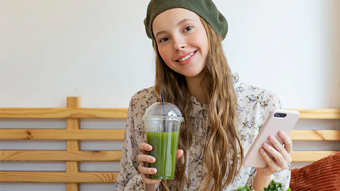 Ein junges Mädchen mit einer grünen Mütze sitzt auf einer Bank. In einer Hand hält sie ihr Handy, in der anderen Hand hält sie einen Plastikbecher mit Strohhalm, in dem ein grüner Saft enthalten ist. Sie lächelt in die Kamera.