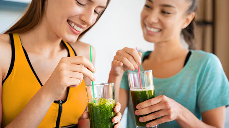 Zwei Frauen stehen nebeneinander und jede von ihnen hat einen grünen Saft in einem Glas in der Hand.