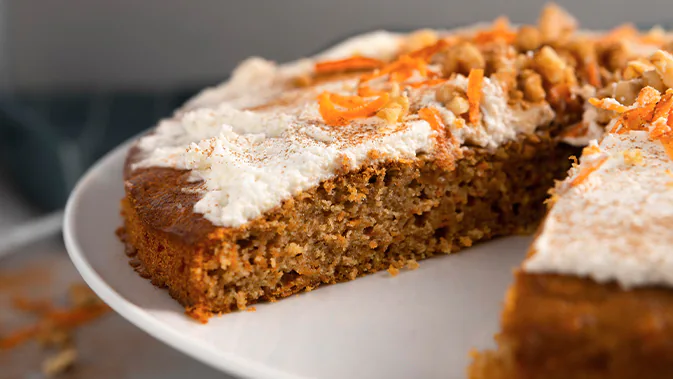 Ein angeschnittener Karottenkuchen auf einem Teller. Man sieht das innere des Kuchens. Auf dem Kuchen ist eine weiße Glasur, sowie Karottenraspeln und Nussstückchen.