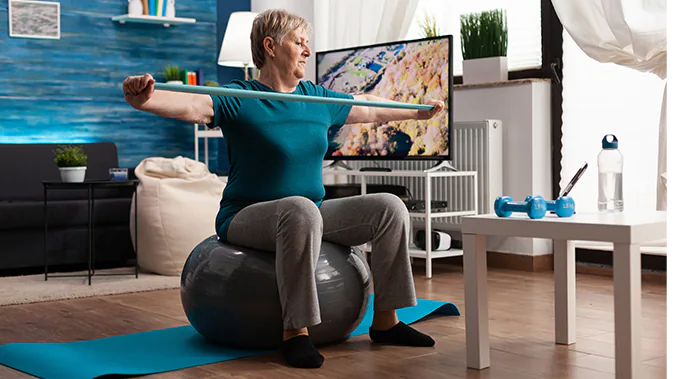 Eine Frau sitzt auf einem Gymnastikball und hält ein Thraband in ihren Händen, welches sie auseinanderzieht und damit Spannung in ihren Armen aufbaut.