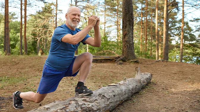 Ein älterer Mann in Sportkleidung ist im Wald und macht an einem liegenden Baumstamm Lunges. Er steht mit einem Fuß auf dem Baumstamm und der andere Fuß steht auf dem Boden.