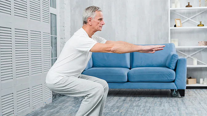 Ein älterer Mann in sportlicher Kleidung führt eine Kniebeuge aus. Er befindet sich im Wohnzimmer vor einem blauen Sofa.