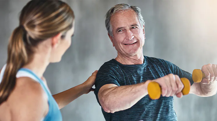 Ein Mann im T-Shirt und mit grauen Haaren hält Kurzhanteln in der Hand und macht eine Übung für die Arme. Eine Frau steht neben ihm und betrachtet seine Übungsausführung.