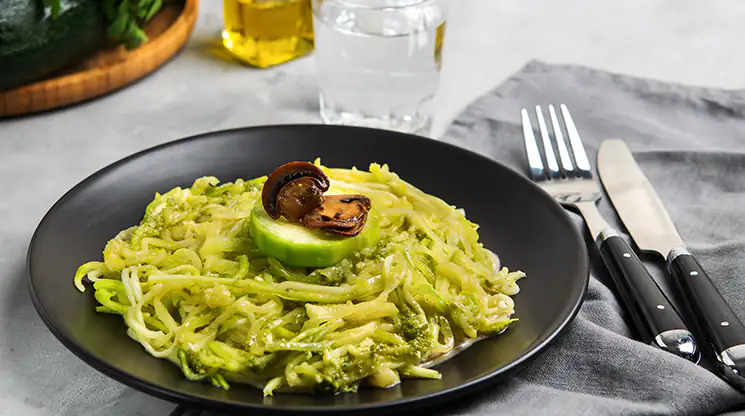 Bei einer Glutenunverträglichkeit ist Gemüse in der Regel immer eine gute Wahl. Zucchinispaghetti garniert mit einem Stück Gurke und Champignons auf einem Teller.