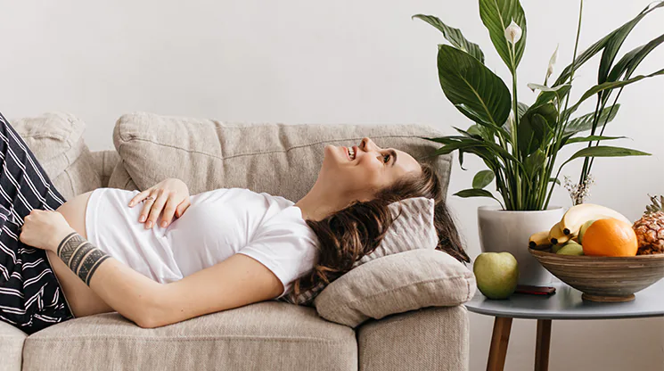 Eine schwangere Frau liegt auf dem Sofa und lacht. Sie hat eine Hand auf ihrem Bauch liegen. Nbene dem Sofa steht ein kleiner Tisch. Auf ihm befindet sich eine Pflanze und eine Obstschale gefüllt mit Orangen, Bananen und Ananas.