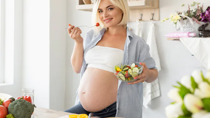 EIne schwangere Frau hält eine Schüssel voll Salat in der einen Hand. Mit der anderen Hand führt sie eine Gabel mit einer Tomate zum Mund. 