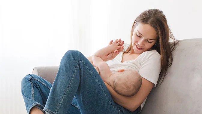 Eine Frau in Jeans und weißem T-Shirt sitzt mit ihrem Baby im Schoß auf dem Sofa. Das Baby wird gerade gestillt und hält seine Hand gegen eine Hand der Mutter.