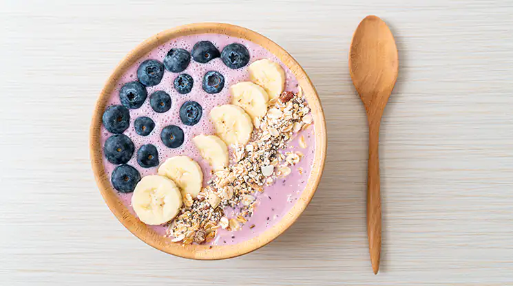 EIne Frühstücksbowl mit einer rosafarbenen Basis, getoppt mit Heidelbeeren, Bananenscheiben und Müsli. Daneben liegt ein Holzlöffel.