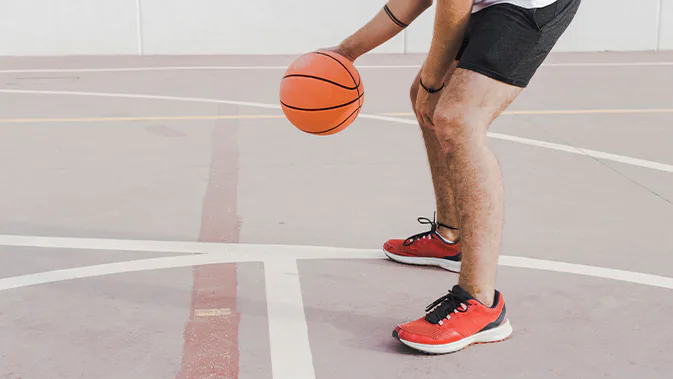 Ein sportlich gekleideter Mann dribbelt einen Basketball. Man sieht den Körper nur ab der Hüfte nach unten , sowie die Arme.
