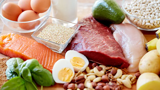 Eine bunte Vielfalt an Lebensmitteln: Eier, Fleisch, Lachs, Nüsse, Kartoffeln, Avocado und Blattspinat liegen nebeneinander auf einem Holzbrett