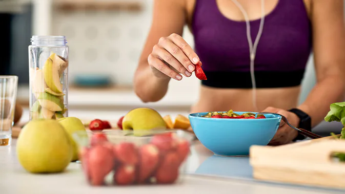 Im Hintergrund ist eine Frau zu sehen, die sich gerade eine gesunde Mahlzeit zubereitet. Im Vordergrund des Bildes steht eine Schale mit Erdbeeren, dahinter ist weiteres Obst zu sehen.