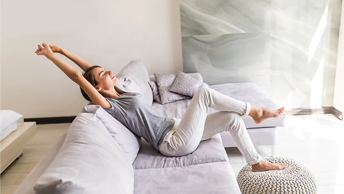 Eine Frau sitzt auf dem Sofa und streckt sich. Sie sieht entspannt und ausgeruht aus.