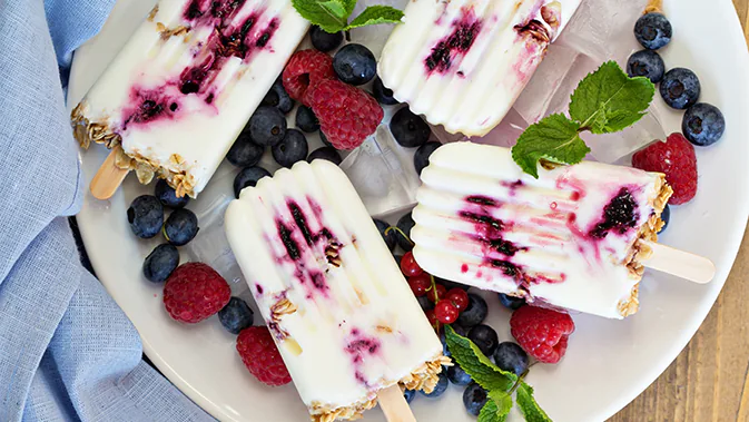 Auf einem Teller liegen 4 Joghurt-Popsicles (Eis am Stil) bestehend aus Joghurt, Beeren und Haferflocken. Rundherum sind Erdbeeren und Heidelbeeren dekoriert.