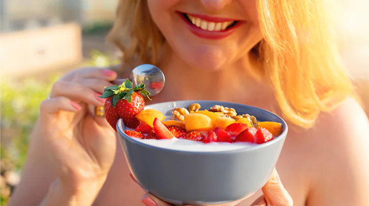 Eine Frau hält eine Schüssel mit Joghurt und Früchten in der Hand, sowie einen Löffel in der anderen Hand. Sie lächelt und freut sich offensichtlich über den leckeren Sommersnack.