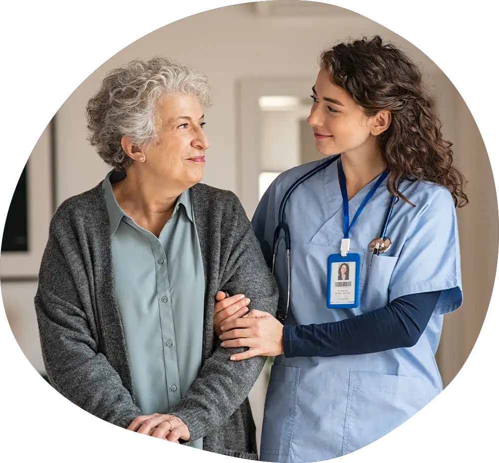Pflegehelferin begleitet pflegebedürftige ältere Frau und lächelt sie an.