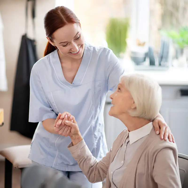 eine Alltagsbegleiterin hält die Hand einer Patientin und die hat die andere Hand auf ihre Schulter gelegt, beide schauen sich vertraut an
