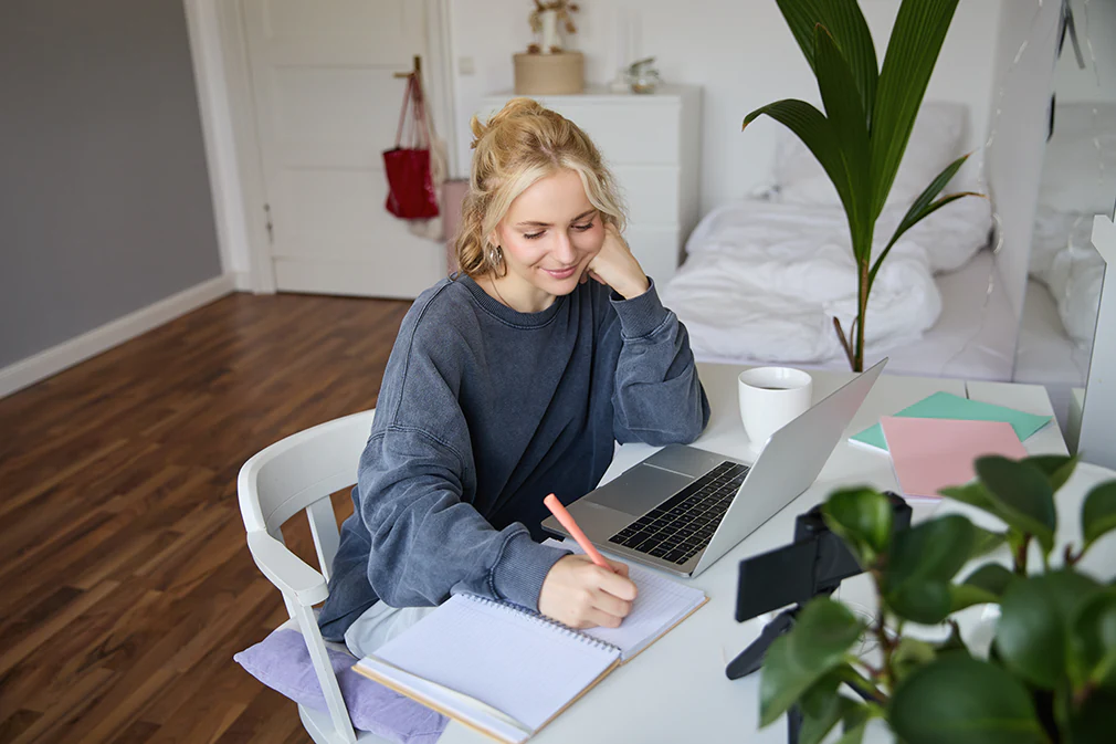 Junge Frau lernt online am Laptop, macht sich Notizen in ein Heft und sitzt entspannt im Schlafzimmer.