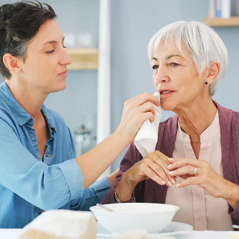 Eine Alltagsbegleiterin tupft einer älteren Patientin den Mundwinkel ab nach dem Essen