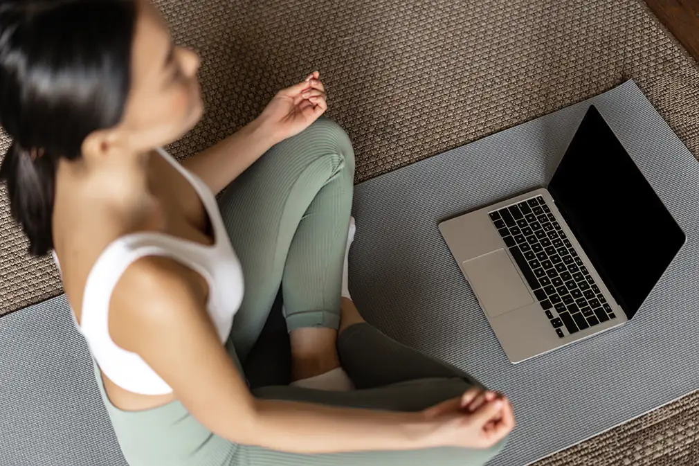 Frau sitzt im Schneidersitz auf Yoga-Matte und vor ihr steht ein Laptop.