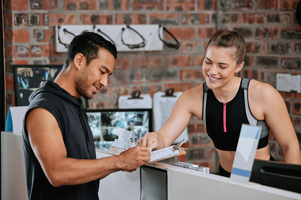 Fitnessstrainerin hilft neuem Mitglied beim Ausfüllen des Anmeldeformulars am Empfang.