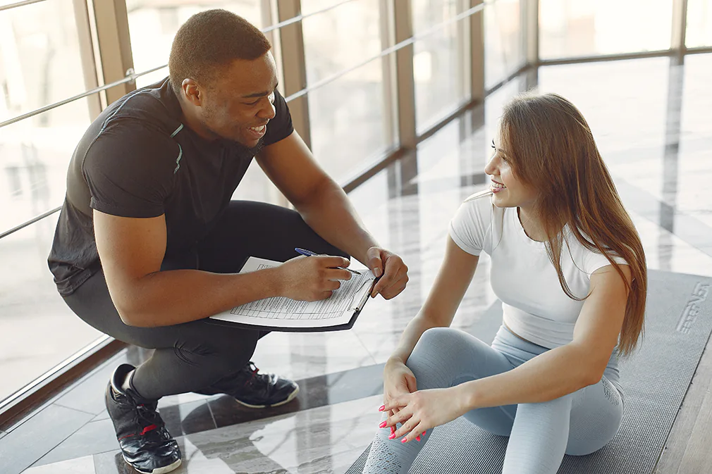 Gesundheitstrainer spricht mit Klientin im Fitnessstudio und bespricht Trainingsplan auf der Matte.