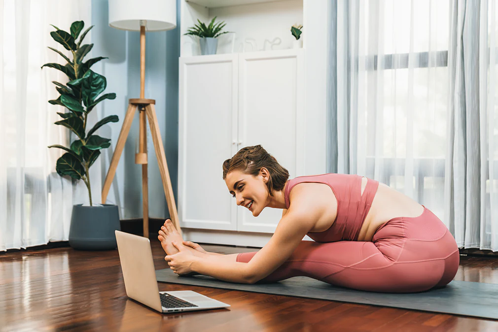 Frau macht Online-Yoga zu Hause und dehnt sich im Sitzen auf der Matte vor dem Laptop.
