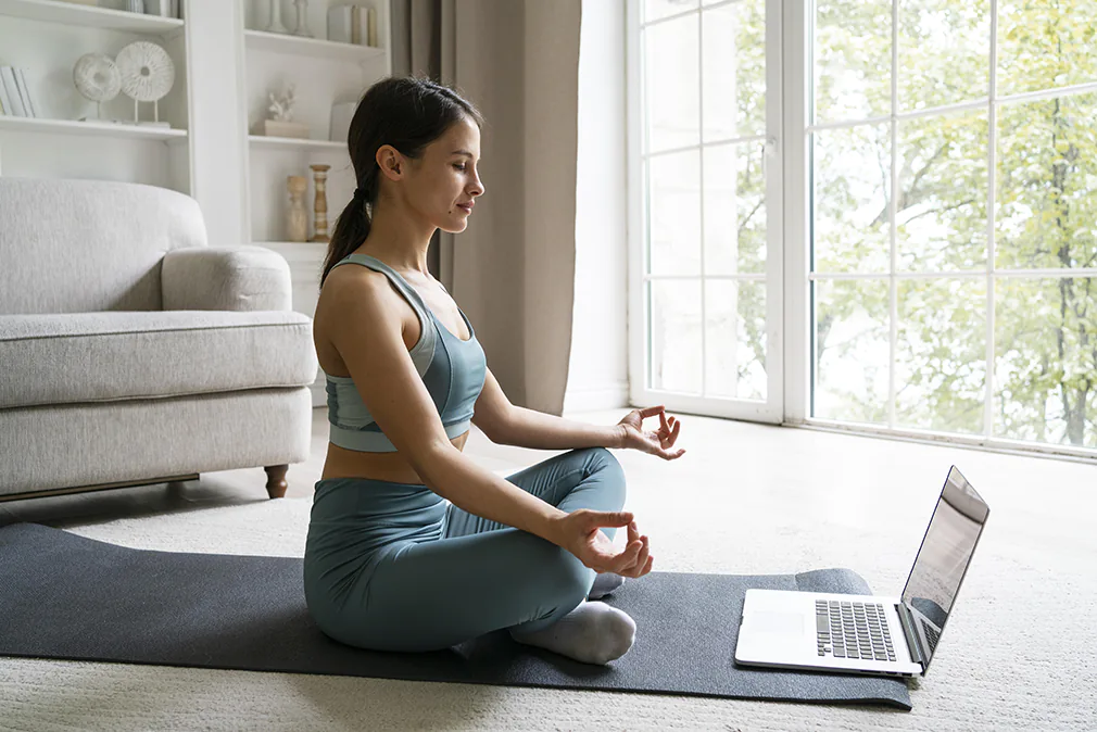 Frau meditiert auf Yogamatte vor dem Laptop und folgt einer Online-Meditation im hellen Wohnzimmer.