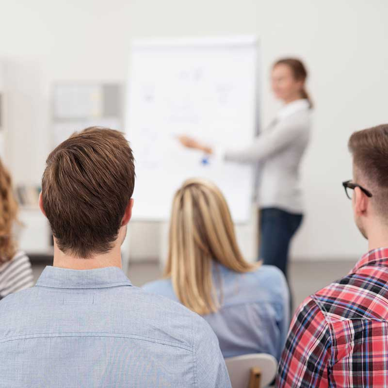Eine Frau führt mit Hilfe eines Flipcharts eine Schulung durch, mehrere Personen hören ihr aufmerksam zu