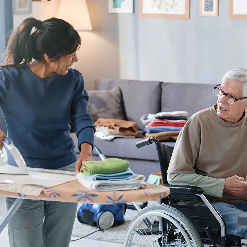 Eine Alltagsbegleiterin bügelt Handtücher eines im Rollstuhl sitzenden älteren Mannes